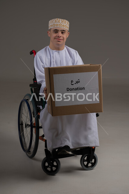 ذوي الإحتياجات الخاصة والهمم, بورتريه لشاب عربي خليجي عماني يرتدي الكمه والدشداشة ينظر الى الكاميرا بتعابير السرور ويحمل بيديه صندوق تبرع، مفهوم مساعدة المحتاجين، خلفية رمادي