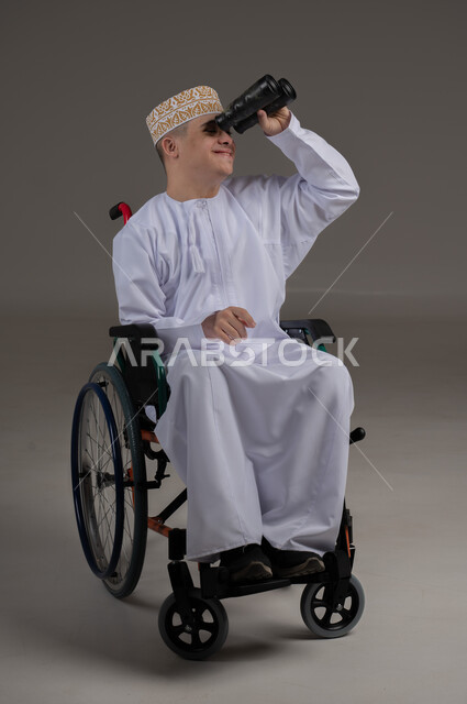 Using binoculars to detect distant objects, people with special needs and disabilities, portrait of an Arab Gulf Omani young man wearing a sleeve and a dishdasha and sitting in a wheelchair, gray background