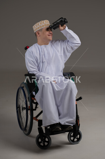 Using binoculars to detect distant objects, people with special needs and disabilities, portrait of an Arab Gulf Omani young man wearing a sleeve and a dishdasha and sitting in a wheelchair, gray background