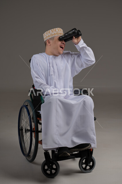 Using binoculars to detect distant objects, people with special needs and disabilities, portrait of an Arab Gulf Omani young man wearing a sleeve and a dishdasha and sitting in a wheelchair, gray background