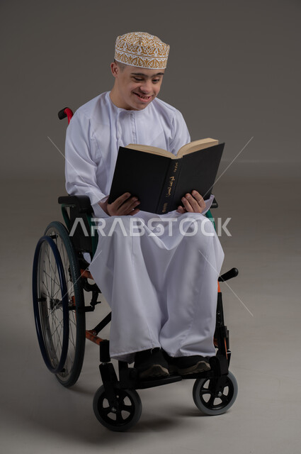 مفهوم القراءة والتعلم والتثقف، ذوي الإحتياجات الخاصة والهمم, بورتريه لشاب عربي خليجي عماني يرتدي الكمه والدشداشة ويجلس على كرسي متحرك ويحمل بيديه كتاب، خلفية رمادي