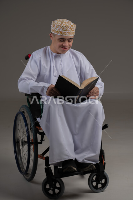 مفهوم القراءة والتعلم والتثقف، ذوي الإحتياجات الخاصة والهمم, بورتريه لشاب عربي خليجي عماني يرتدي الكمه والدشداشة ويجلس على كرسي متحرك ويحمل بيديه كتاب، خلفية رمادي