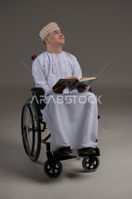 مفهوم قراءة القران وحفظه وتجويده، ذوي الإحتياجات الخاصة والهمم, بورتريه لشاب عربي خليجي عماني يرتدي الكمه والدشداشة يجلس على كرسي متحرك ويحمل بيديه المصحف الشريف، خلفية رمادي
