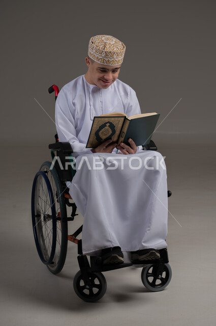مفهوم قراءة القران وحفظه وتجويده، ذوي الإحتياجات الخاصة والهمم, بورتريه لشاب عربي خليجي عماني يرتدي الكمه والدشداشة يجلس على كرسي متحرك ويحمل بيديه المصحف الشريف، خلفية رمادي