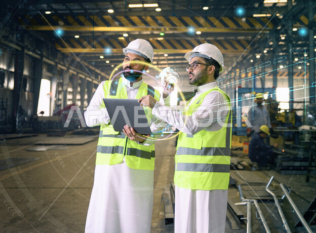 مهندسان عربيان خليجيان سعوديان يرتديان الخوذة والسترة الخاصة بالعمل داخل مصنع الحديد ،استخدام الحاسوب المحمول لفحص جودة الحديد، استخدام تقنية الهولجرام ثلاثية الابعاد