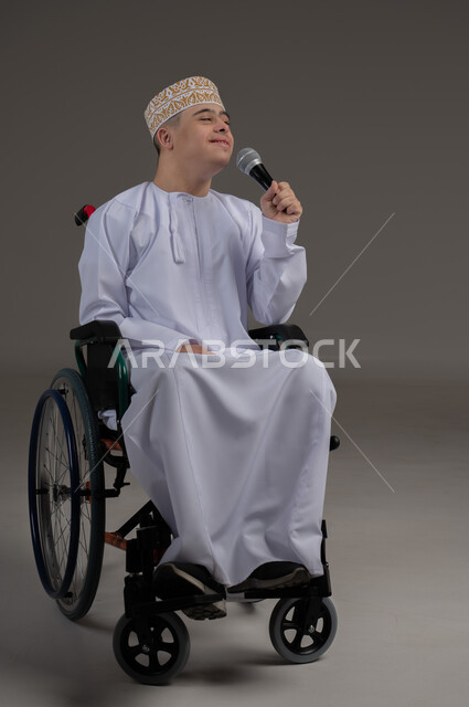 مفهوم الانسجام بالغناء، ذوي الإحتياجات الخاصة والهمم, بورتريه لشاب عربي خليجي عماني يرتدي الكمه والدشداشة يجلس على كرسي متحرك ويحمل بيده مايكروفون، خلفية رمادي