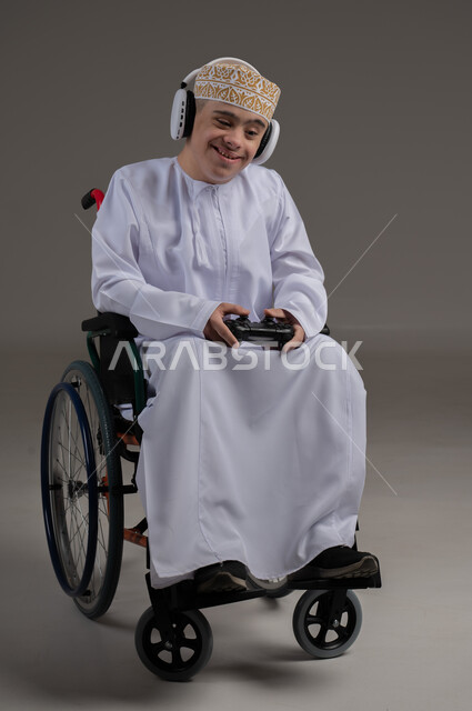 قضاء اوقات ممتعة اثناء اللعب، مفهوم الترفيه والتسلية, ذوي الإحتياجات الخاصة والهمم, بورتريه لشاب عربي خليجي عماني يرتدي الكمه والدشداشة يجلس على كرسي متحرك ويضع سماعات الرأس، خلفية رمادي