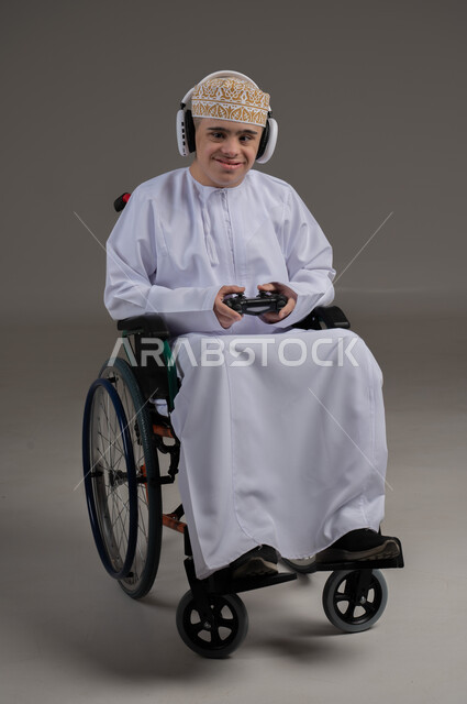 قضاء اوقات ممتعة اثناء اللعب، مفهوم الترفيه والتسلية, ذوي الإحتياجات الخاصة والهمم, بورتريه لشاب عربي خليجي عماني يرتدي الكمه والدشداشة يجلس على كرسي متحرك ويضع سماعات الرأس، خلفية رمادي
