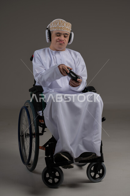قضاء اوقات ممتعة اثناء اللعب، مفهوم الترفيه والتسلية, ذوي الإحتياجات الخاصة والهمم, بورتريه لشاب عربي خليجي عماني يرتدي الكمه والدشداشة يجلس على كرسي متحرك ويضع سماعات الرأس، خلفية رمادي