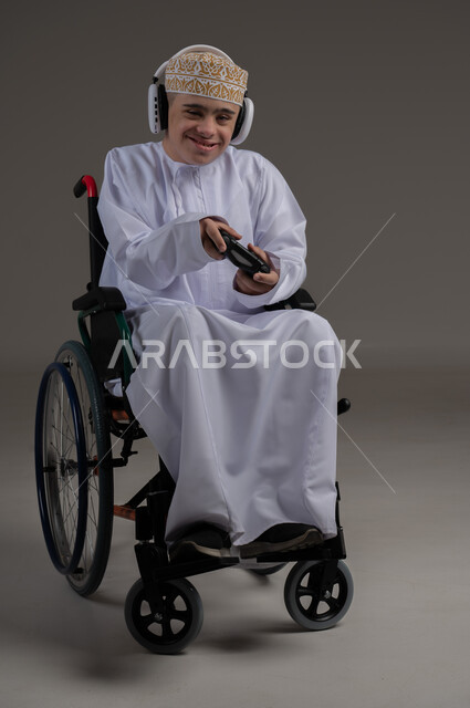 Spending fun times while playing, the concept of entertainment and amusement, people with special needs and concerns, portrait of an Arab Gulf Omani young man wearing a sleeve and a dishdasha, sitting in a wheelchair and wearing headphones, gray background