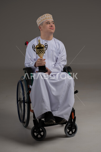 مفهوم المسابقات والربح, ذوي الإحتياجات الخاصة والهمم, بورتريه لشاب عربي خليجي عماني يرتدي الكمه والدشداشة يجلس على كرسي متحرك ويحمل بيده كأس البطولة، خلفية رمادي