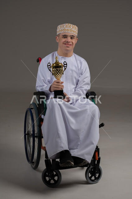 مفهوم المسابقات والربح, ذوي الإحتياجات الخاصة والهمم, بورتريه لشاب عربي خليجي عماني يرتدي الكمه والدشداشة يجلس على كرسي متحرك ويحمل بيده كأس البطولة، خلفية رمادي