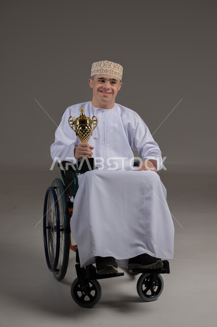 مفهوم المسابقات والربح, ذوي الإحتياجات الخاصة والهمم, بورتريه لشاب عربي خليجي عماني يرتدي الكمه والدشداشة يجلس على كرسي متحرك ويحمل بيده كأس البطولة، خلفية رمادي
