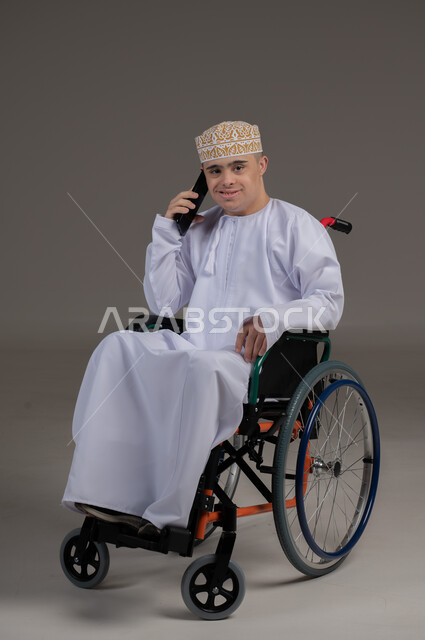 مفهوم التواصل مع الاهل والاصدقاء, ذوي الإحتياجات الخاصة والهمم, بورتريه لشاب عربي خليجي عماني يرتدي الكمه والدشداشة يجلس على كرسي متحرك ويحمل بيده هاتف محمول بايماءات التحدث مع شخص، خلفية رمادي