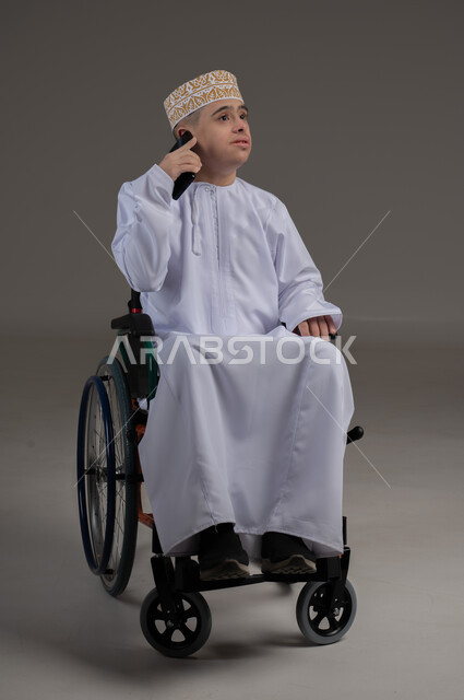 مفهوم التواصل مع الاهل والاصدقاء, ذوي الإحتياجات الخاصة والهمم, بورتريه لشاب عربي خليجي عماني يرتدي الكمه والدشداشة يجلس على كرسي متحرك ويحمل بيده هاتف محمول بايماءات التحدث مع شخص، خلفية رمادي