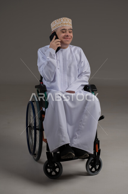 مفهوم التواصل مع الاهل والاصدقاء, ذوي الإحتياجات الخاصة والهمم, بورتريه لشاب عربي خليجي عماني يرتدي الكمه والدشداشة يجلس على كرسي متحرك ويحمل بيده هاتف محمول بايماءات التحدث مع شخص، خلفية رمادي