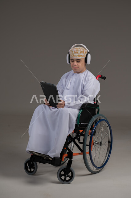 مفهوم التواصل مع الاهل والاصدقاء, ذوي الإحتياجات الخاصة والهمم, بورتريه لشاب عربي خليجي عماني يرتدي الكمه والدشداشة يجلس على كرسي متحرك يضع سماعات الراس ويحمل بيده جهاز تابليت  بايماءات التحدث مع شخص، خلفية رمادي