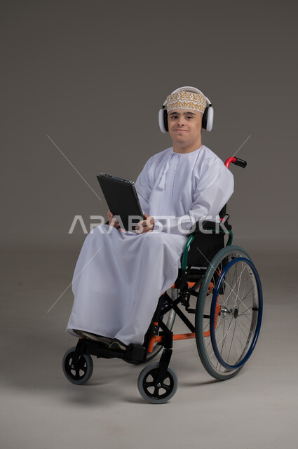 مفهوم التواصل مع الاهل والاصدقاء, ذوي الإحتياجات الخاصة والهمم, بورتريه لشاب عربي خليجي عماني يرتدي الكمه والدشداشة يجلس على كرسي متحرك يضع سماعات الراس ويحمل بيده جهاز تابليت  بايماءات التحدث مع شخص، خلفية رمادي