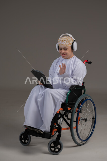 مفهوم التواصل مع الاهل والاصدقاء, ذوي الإحتياجات الخاصة والهمم, بورتريه لشاب عربي خليجي عماني يرتدي الكمه والدشداشة يجلس على كرسي متحرك يضع سماعات الراس ويحمل بيده جهاز تابليت  بايماءات التحدث مع شخص، خلفية رمادي