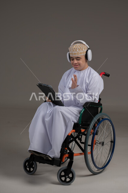 مفهوم التواصل مع الاهل والاصدقاء, ذوي الإحتياجات الخاصة والهمم, بورتريه لشاب عربي خليجي عماني يرتدي الكمه والدشداشة يجلس على كرسي متحرك يضع سماعات الراس ويحمل بيده جهاز تابليت  بايماءات التحدث مع شخص، خلفية رمادي