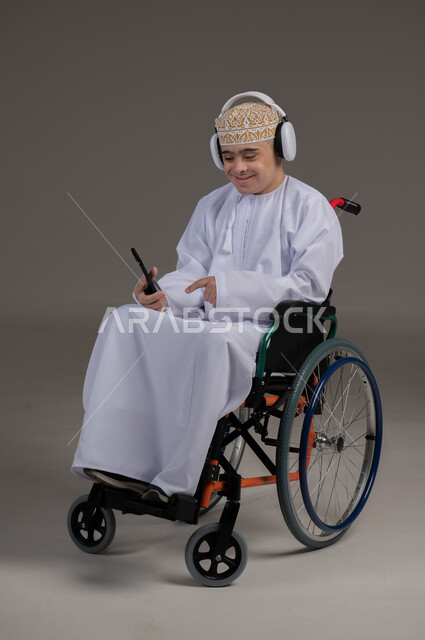 مفهوم التواصل مع الاهل والاصدقاء, ذوي الإحتياجات الخاصة والهمم, بورتريه لشاب عربي خليجي عماني يرتدي الكمه والدشداشة يجلس على كرسي متحرك يضع سماعات الراس ويمسك الهاتف المحمول, خلفية رمادي