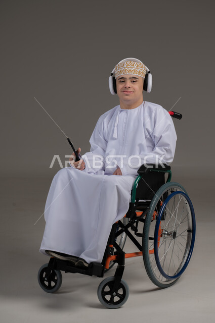 مفهوم التواصل مع الاهل والاصدقاء, ذوي الإحتياجات الخاصة والهمم, بورتريه لشاب عربي خليجي عماني يرتدي الكمه والدشداشة يجلس على كرسي متحرك يضع سماعات الراس ويمسك الهاتف المحمول, خلفية رمادي