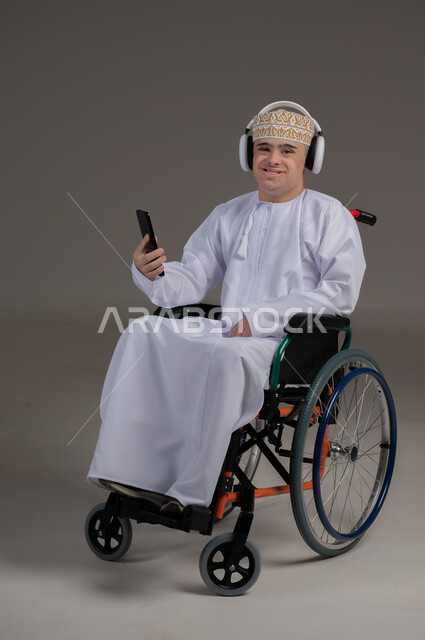 مفهوم التواصل مع الاهل والاصدقاء, ذوي الإحتياجات الخاصة والهمم, بورتريه لشاب عربي خليجي عماني يرتدي الكمه والدشداشة يجلس على كرسي متحرك يضع سماعات الراس ويمسك الهاتف المحمول, خلفية رمادي