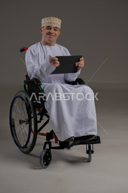 ذوي الإحتياجات الخاصة والهمم, مفهوم اللعب والتسلية , بورتريه لشاب عربي خليجي عماني يرتدي الكمه والدشداشة يجلس على كرسي متحرك ويحمل بيديه جهاز تابليت, خلفية رمادي