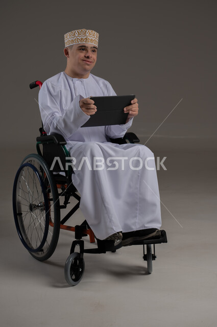 ذوي الإحتياجات الخاصة والهمم, مفهوم اللعب والتسلية , بورتريه لشاب عربي خليجي عماني يرتدي الكمه والدشداشة يجلس على كرسي متحرك ويحمل بيديه جهاز تابليت, خلفية رمادي