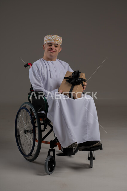 ذوي الإحتياجات الخاصة والهمم, مفهوم الاحتفال والهداية, بورتريه لشاب عربي خليجي عماني يرتدي الكمه والدشداشة يجلس على كرسي متحرك ويحمل بيده هدية, خلفية رمادي