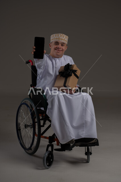 ذوي الإحتياجات الخاصة والهمم, مفهوم الاحتفال والهداية, بورتريه لشاب عربي خليجي عماني يرتدي الكمه والدشداشة يجلس على كرسي متحرك ويحمل بيده هدية, خلفية رمادي