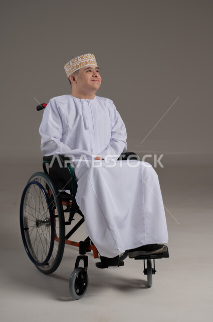 People with special needs and concerns, gestures of happiness and pleasure, portrait of an Arab Gulf Omani young man wearing a sleeve and a dishdasha sitting in a wheelchair, gray background