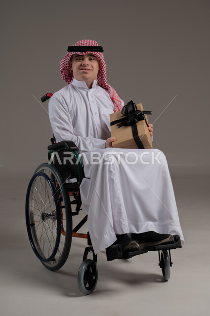ذوي الإحتياجات الخاصة والهمم, مفهوم الهداية والاعياد, بورتريه لشاب عربي خليجي سعودي يرتدي الثوب والشماغ يجلس على كرسي متحرك ويحمل بيديه هدية, خلفية رمادي