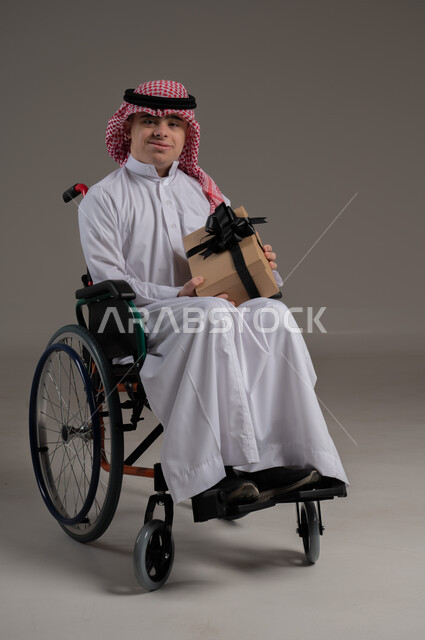 ذوي الإحتياجات الخاصة والهمم, مفهوم الهداية والاعياد, بورتريه لشاب عربي خليجي سعودي يرتدي الثوب والشماغ يجلس على كرسي متحرك ويحمل بيديه هدية, خلفية رمادي