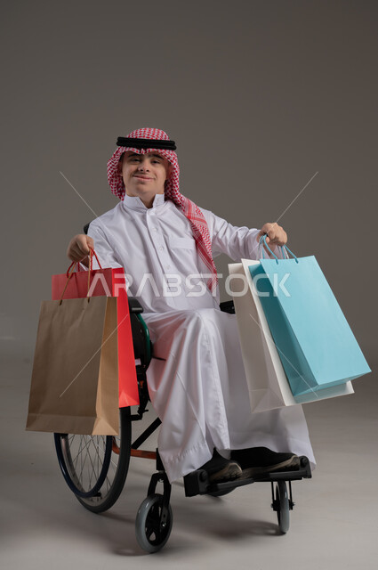 مفهوم التسوق والتبضع, ذوي الإحتياجات الخاصة والهمم, بورتريه لشاب عربي خليجي سعودي يرتدي الثوب والشماغ يجلس على كرسي متحرك ويحمل بيديه اكياس, خلفية رمادي
