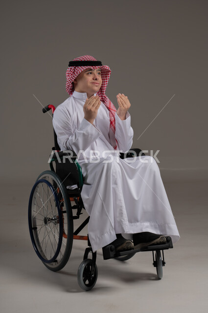 مفهوم طلب الحاجة من الله, ذوي الإحتياجات الخاصة والهمم, بورتريه لشاب عربي خليجي سعودي يرتدي الثوب والشماغ يجلس على كرسي متحرك, رفع اليدين بايماءات الدعاء, خلفية رمادي
