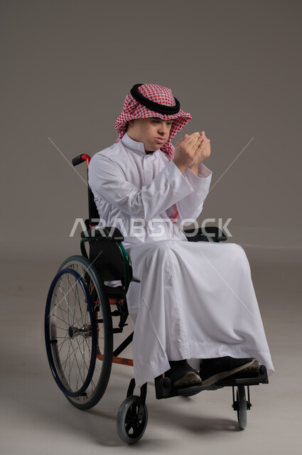 مفهوم طلب الحاجة من الله, ذوي الإحتياجات الخاصة والهمم, بورتريه لشاب عربي خليجي سعودي يرتدي الثوب والشماغ يجلس على كرسي متحرك, رفع اليدين بايماءات الدعاء, خلفية رمادي