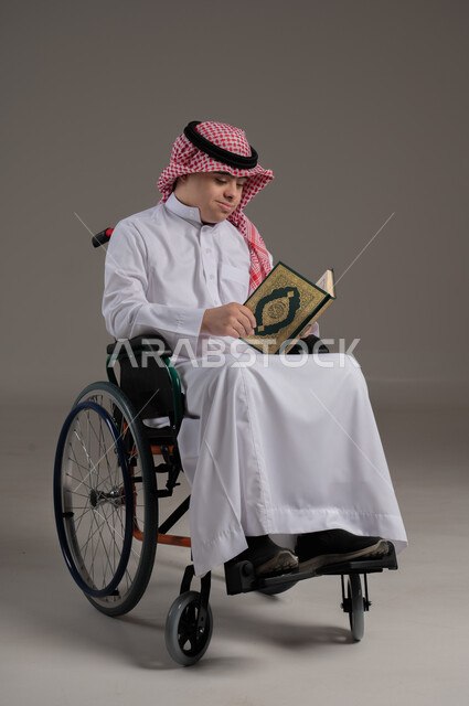مفهوم حفظ القران وتلاوته, ذوي الإحتياجات الخاصة والهمم, بورتريه لشاب عربي خليجي سعودي يرتدي الثوب والشماغ يجلس على كرسي متحرك ويمسك بيديه المصحف الشريف, خلفية رمادي