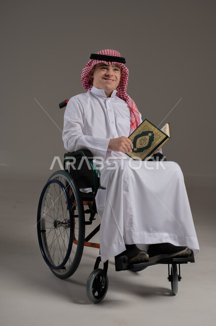 مفهوم حفظ القران وتلاوته, ذوي الإحتياجات الخاصة والهمم, بورتريه لشاب عربي خليجي سعودي يرتدي الثوب والشماغ يجلس على كرسي متحرك ويمسك بيديه المصحف الشريف, خلفية رمادي