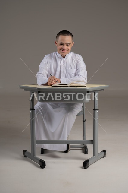 Integrating people with special needs in Omani schools, developing curricula and educational materials, portrait of a young Gulf Arab Omani man with Down syndrome wearing a dishdasha sitting at a study table reviewing lessons, full-length body image, gray background