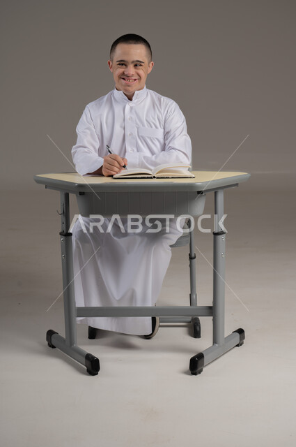تطوير المناهج والمواد التعليمية، دمج ذوي الاحتياجات الخاصة في المدارس العمانية، بورتريه لشاب عربي خليجي عماني مصاب بمتلازمة داون يرتدي الدشداشة يجلس الى طاولة الدراسة يقوم بمراجعه الدروس ، صورة الجسم بالطول الكامل، خلفية رمادية