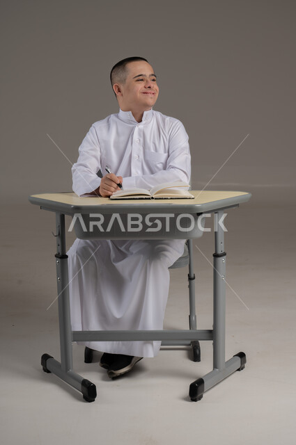 Gestures of distraction and lack of concentration, integration of people with special needs in Omani schools, development of curricula and educational materials, portrait of a young Gulf Arab Omani man with Down syndrome wearing a dishdasha sitting at a study table reviewing lessons, full-length body image, gray background