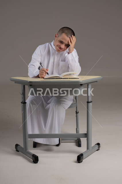 دمج ذوي الاحتياجات الخاصة في المدارس العمانية، تطوير المناهج والمواد التعليمية، بورتريه لشاب عربي خليجي عماني مصاب بمتلازمة داون يرتدي الدشداشة يجلس الى طاولة الدراسة يقوم بمراجعه الدروس ، صورة الجسم بالطول الكامل، خلفية رمادية