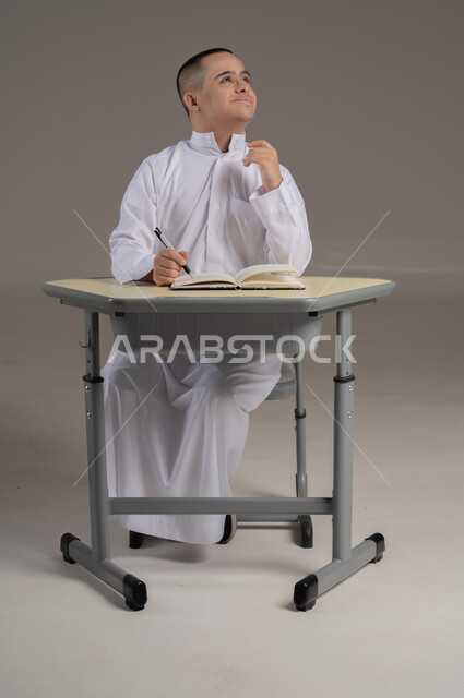 تطوير المناهج والمواد التعليمية، دمج ذوي الاحتياجات الخاصة في المدارس العمانية، بورتريه لشاب عربي خليجي عماني مصاب بمتلازمة داون يرتدي الدشداشة يجلس الى طاولة الدراسة يقوم بمراجعه الدروس ، صورة الجسم بالطول الكامل، خلفية رمادية