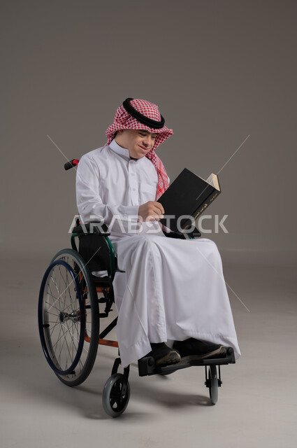 مفهوم القراءة والمطالعة والتثقف, ذوي الإحتياجات الخاصة والهمم, بورتريه لشاب عربي خليجي سعودي يرتدي الثوب والشماغ يجلس على كرسي متحرك ويمسك بيديه كتاب, خلفية رمادي