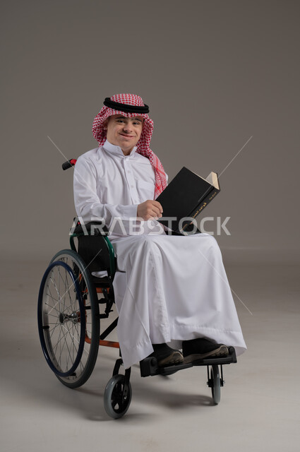 مفهوم القراءة والمطالعة والتثقف, ذوي الإحتياجات الخاصة والهمم, بورتريه لشاب عربي خليجي سعودي يرتدي الثوب والشماغ يجلس على كرسي متحرك ويمسك بيديه كتاب, خلفية رمادي