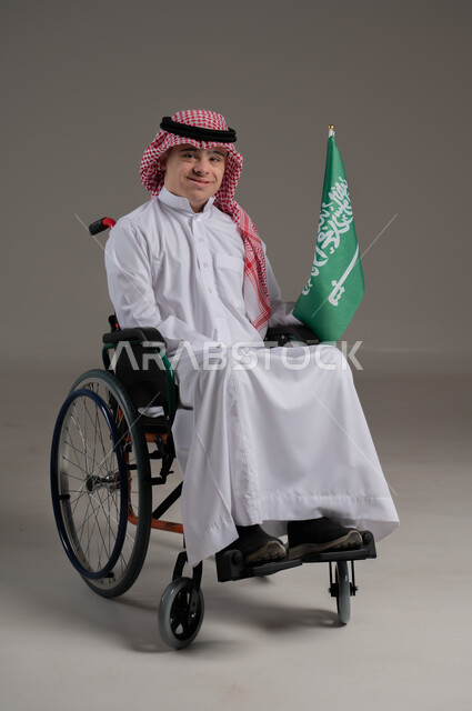 مفهوم حب الوطن والانتماء اليه, ذوي الإحتياجات الخاصة والهمم, بورتريه لشاب عربي خليجي سعودي يرتدي الثوب والشماغ يجلس على كرسي متحرك ويمسك بيده علم المملكة العربية السعودية, خلفية رمادي