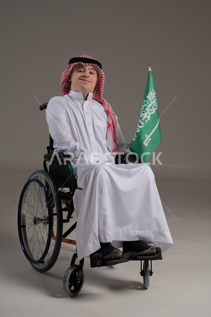 مفهوم حب الوطن والانتماء اليه, ذوي الإحتياجات الخاصة والهمم, بورتريه لشاب عربي خليجي سعودي يرتدي الثوب والشماغ يجلس على كرسي متحرك ويمسك بيده علم المملكة العربية السعودية, خلفية رمادي