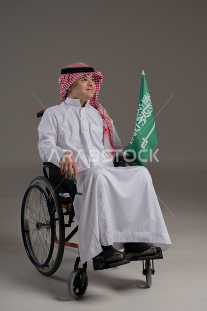 مفهوم حب الوطن والانتماء اليه, ذوي الإحتياجات الخاصة والهمم, بورتريه لشاب عربي خليجي سعودي يرتدي الثوب والشماغ يجلس على كرسي متحرك ويمسك بيده علم المملكة العربية السعودية, خلفية رمادي