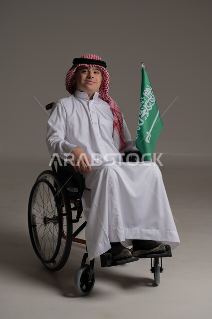 مفهوم حب الوطن والانتماء اليه, ذوي الإحتياجات الخاصة والهمم, بورتريه لشاب عربي خليجي سعودي يرتدي الثوب والشماغ يجلس على كرسي متحرك ويمسك بيده علم المملكة العربية السعودية, خلفية رمادي